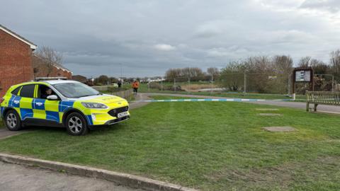A section of a park has been cordoned off with blue and white police take. A police car is parked on the green in the left-hand-corner of the park. Trees and fencing around more parkland are in the background.