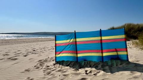 A sandy beach scene on a clear sunny day. A windbreak has been placed on the sand but there are no people in sight.