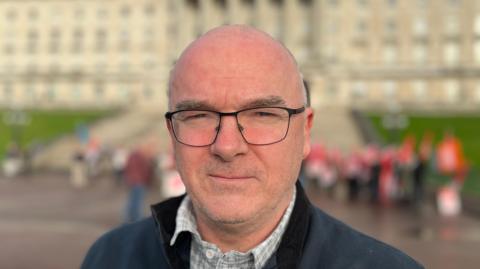 A bald man with black glasses and clear frames. 
He is wearing a navy sweatshirt and a grey shirt. 
The background is blurred but a white building and feint pictures of protesters can be pictured standing behind him