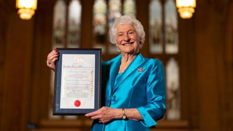 Lady Mary is standing, smiling at the camera and holding her award. She is wearing a blue dress with a silver brooch, a watch and has red, painted nails. 