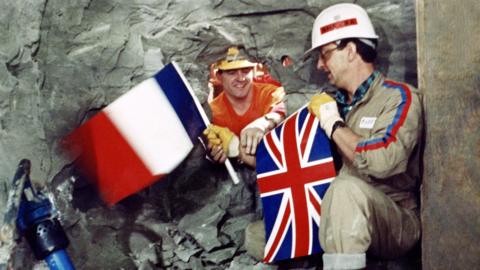 Tunnel workers Philippe Cozette from France (R) and Graham Fagg from England shake hands while holding national flags on December 01, 1990, during the historic breakthrough in the Channel Tunnel.