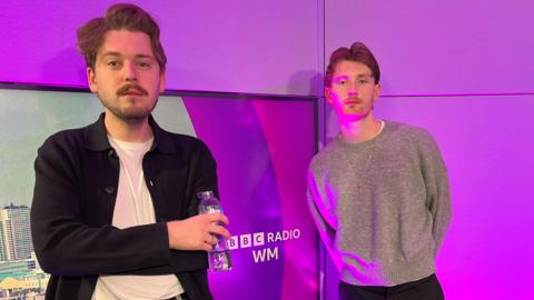 Two men standing next to each other against a purple background with the words "BBC Radio WM" on it. The man on the left has short brown hair and a moustache and is wearing a white T-shirt and black cardigan. The man on the right has short brown hair and is wearing a grey jumper. 