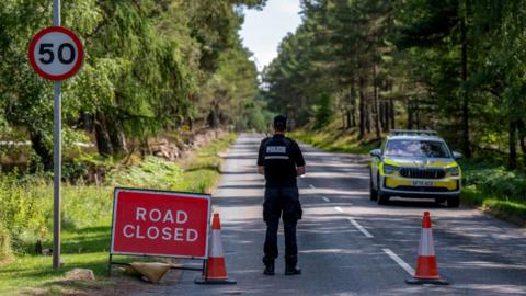 Closed road. A police officer is standing on a road with his back to the camera. He is next to a red and white Road Closed sign and two orange and white traffic cones. There is a police car further down the road
