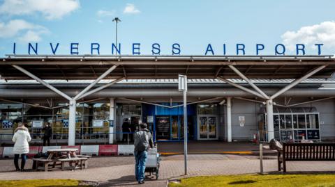 An exterior view of Inverness Airport showing its entrance with two passengers outside it. Above the entrance is a sign spelling out: "Inverness Airport" in capital letters.