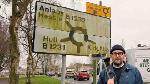 A man on the side of a road holding a large brush, of the type used by window cleaners, with a large, white, dirty road sign behind him. He is wearing a blue jacket and beanie hat. A white van is parked by the side of the road. On the back it says, "we clean stuff really good!" The road sign is covered in green moss. It has a roundabout symbol on it, with exits pointing to Anlaby, Hessle, B1232, Hull B1231 and Kirk Ella.
