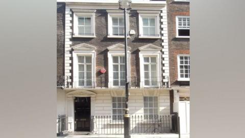 Front view of a 18th Century brick townhouse in London. It has a black front door and appears to be three storeys tall. There is a traditional black lamppost on the pavement outside