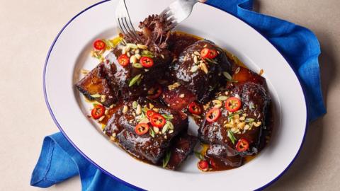 A white oval bowl of short ribs in a glossy sauce topped with red chilli slices and peanuts, on a beige background