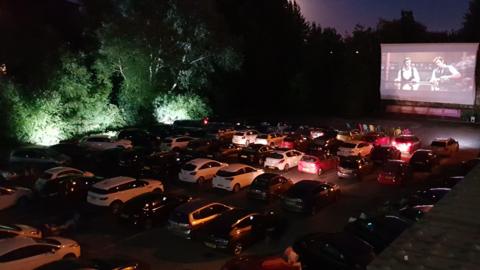 Cars are parked facing a large outdoor cinema screen in the dark. 