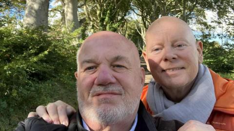 David Letherman and Jim Cunningham pose in a selfie taken by Letherman, who is at the front wearing a black jacket. Cunningham is behind him with his hands on Letherman's shoulders. He is wearing an orange padded jacket and grey scarf. Both men are bald, Letherman has a short grey beard. They are standing in a country lane with hedgerows and trees on each side.