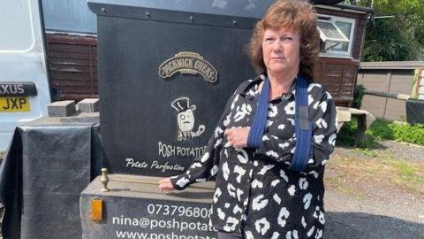 Nina Kelly looking at the camera. She has curly brown hair and is wearing a black shirt with white abstract prints on it, and she has her arm in a blue sling. She is standing in front of her black potato oven. 
