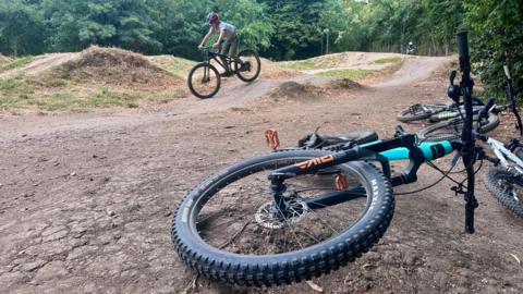 A cyclist wearing a helmet rides a bike over a dirt track surrounded by trees. In the foreground, two bikes lie on the ground beside the trail, with one wheel prominently visible front and centre.