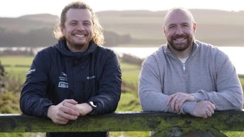 Jonny Snape, nature recovery ranger, and Matthew Squires, from The Squires Foundation. They are outdoors in an area of open countryside, with a lake in the background. They are both leaning on a gate or fence and smiling at the camera. Mr Snape is wearing a dark outdoor jacket, he has long blond curly hair tied back and a short beard. Mr Squires is bald with a trimmed brown beard. He is wearing a grey jumper.