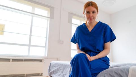 Stacey Dooley in scrubs sitting on a ward bed