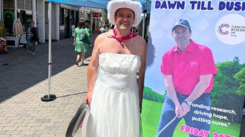A middle-aged man in a white wedding dress, with a fluffy white cowboy hat and a pink neckerchief stands next to a roller banner with a picture of himself playing golf in a pink polo shirt. Text reads: Olly's Golf Challenge Dawn Till Dusk. Cancer Research UK.