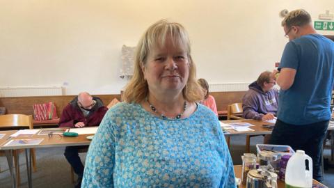 A woman in a patterned blue top stands facing the camera, with other people behind her sat and standing. There are tables set up in a line and a plain, light-coloured wall in the background.