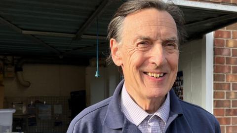Mr Heathcock is standing outside the garage/ shop and smiling. He is wearing a blue collar RNLI top and a blue and white shirt underneath. He has shot brown and grey hair. 