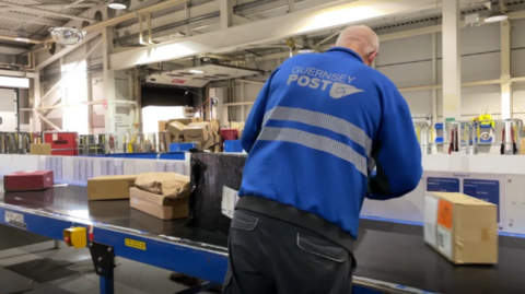 A man wearing a Guernsey Post jacket putting parcels on the moving conveyor belt in the warehouse.