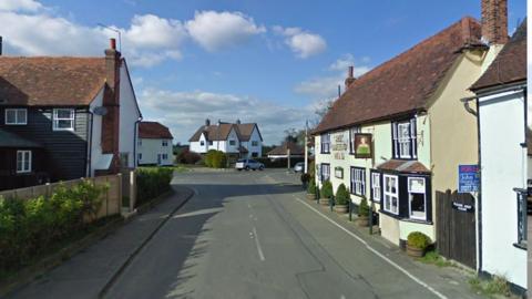 Queen Street, Fyfield, with the Queen's Head pub on the right side. It is an old building with a red tiled roof and chimneys, and a sign of a Queen wearing green on the wall. There are smart houses on the roads around.