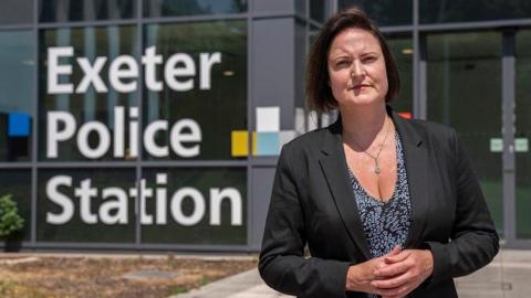 Alison Hernandez is smiling as she stands outside Exeter police station on a sunny day. She has her hands folded in front of her and is wearing a black and white printed top and black jacket. She has dark hair cut into a bob. Behind her is the police station with its name written in large letters in the window/