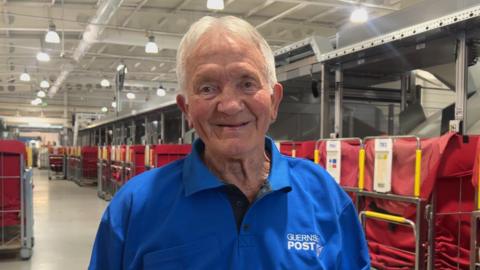 The image shows a man with short white hair smiling at the camera. He is wearing a blue polo shirt that has a "Guernsey Post" logo on it. In the background, is a postage sorting warehouse.