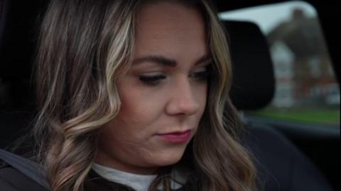 A woman with dark brown and blonde hair looking down. She is sitting in a car and has a seatbelt on