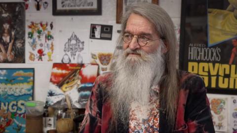 Rob Davies in his shop. He is sitting in front of a wall decorated with eclectic, colourful film posters. He is looking away from the camera. He has long white hair and a long white beard. He is wearing round-rimmed glasses.