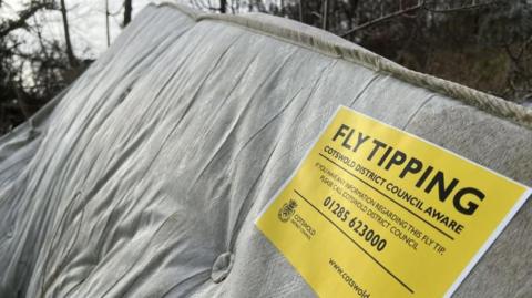 An old mattress dumped near some trees. It has a yellow fly-tipping sticker on it from the council that says 'Fly tipping. Cotswold District Council Aware'. The sticker appeals for anyone with information about the fly-tip to contact them. 