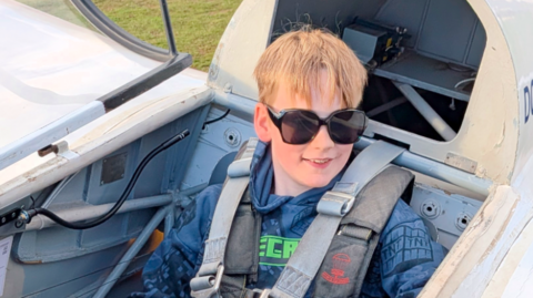 Monty smiling for a photo in an airplane on a field with a seatbelt on.