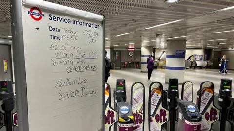 A sign beside some Tube gate barriers in a station says there are severe delays on Victoria and Northern lines. People are in the background