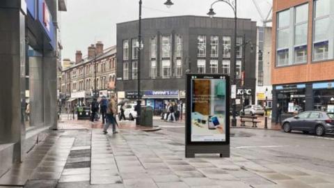 Mock-up image of proposed digital street advertising on wide pavement in shopping street with a KFC restaurant in the background next to a shop with a blue sign and white letters reading Davina's