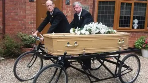 Two men in front of a house with brown windows, on a tandem bike that carries a coffin. 