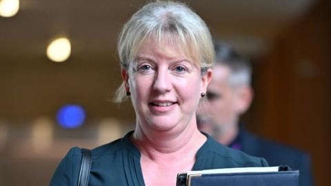 Shona Robison, wearing a blue top, carrying a folder and looking at the camera in the Scottish Parliament 