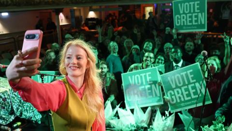 Hannah Spencer doing a selfie with Green Party supporters in the background with placards