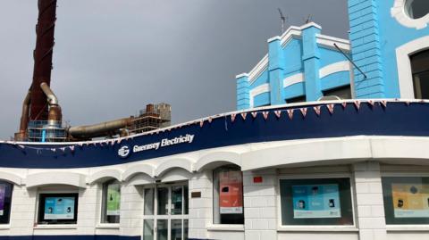 The exterior view of Guernsey Electricity's shop, with a chimney in the left of the image and a blue building rising up above it. 