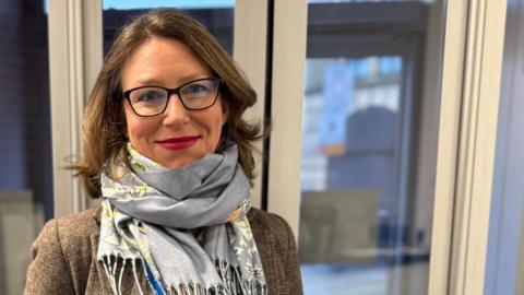 A woman with glasses, in a brown jacket, white shirt and blue scarf stands in a reception room smiling at the camera. 