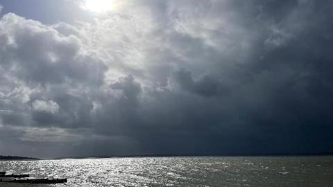 A bright, white sun shines through a break in dark grey clouds above a glistening sea in this dramatic seascape at Hill Head Beach