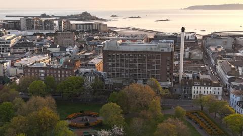 This is an elevated view of St Helier showing a large public park in the foreground, with trees and flower beds. The General Hospital is in view behind and other buildings and the sea are beyond. The coastline, harbour and sea stretch into the distance under warm evening light, with Elizabeth Castle visible on a tidal island offshore.