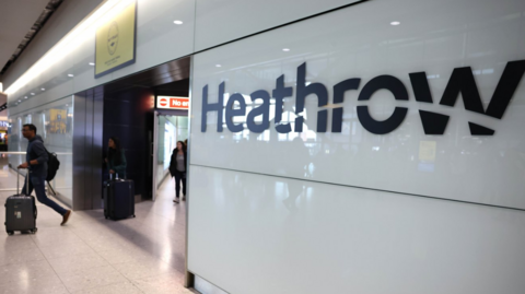 A wide shot of a sign at Heathrow airport which reads 'Heathrow'. Three people are waling through a door into the airport's arrivals hall.