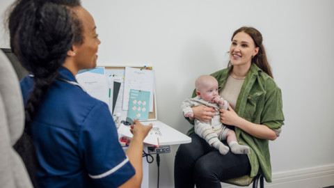 A mother with a baby on her knee meets a healthcare professional in blue scrubs.