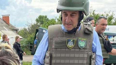 Brooks Newmark wearing protective army clothing with two female Ukrainian refugees just at the corner of the shot. He is standing outside looking down out of shot