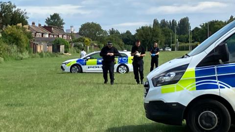 Three police officers dressed in black are walking towards the camera. A police van is parked in the foreground with a police car in the background.