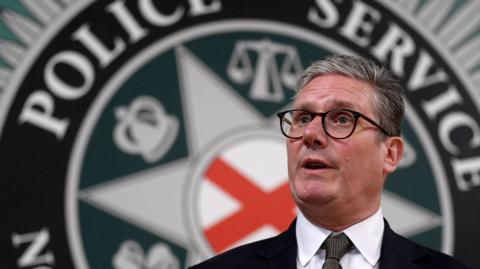 A man with dark grey hair is speaking to someone off-camera. He's standing in front of a blurred PSNI badge and is wearing dark rimmed glasses, a dark-coloured suit and white shirt. 