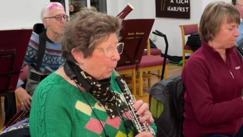 A woman wearing a green jumper is playing oboe with an orchestra