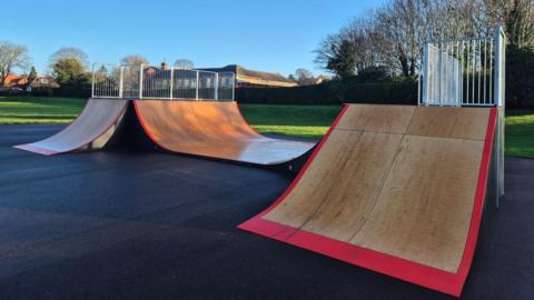 Part of a skatepark on black asphalt. There is a wooden ramp with red outer border and metal railings at the top. To the left of the photo are two other ramps, one broad and one narrow. A green field, trees and houses can be seen in the background.