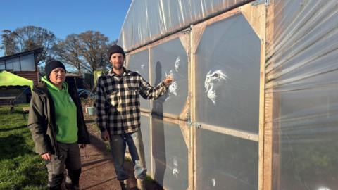 A woman wearing a black woolly hat, green hoodie and a black coat and trousers stands with a man wearing a black woolly hat, a black and white checked shirt and blue jeans. The man rests his hand on the ripped plastic tunnel to his left. 
