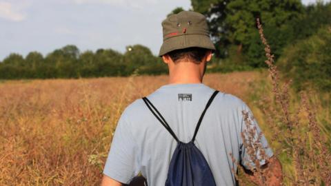 The back of a man walking through a field. He is wearing a green bucket hat and a light blue shirt and is carrying a navy drawstring bag.