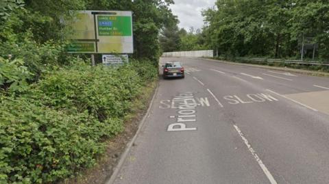 A road with four lanes. A black car is driving away from the camera in the left-hand lane. There are trees and bushes on both sides of the road and a road sign on the left, showing directions to Leeds, the Humber Bridge and Goole.