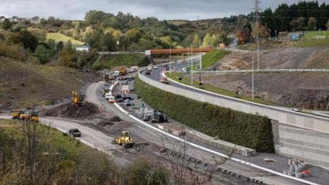 Construction work underway on a split-level dual carriageway, with yellow construction vehicles on the road. Road verges have been partially landscaped.