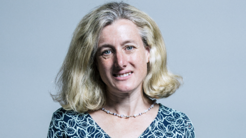 Woman with short blonde hair and a patterned blue top and beaded necklace stands infront of grey background