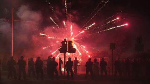 right fireworks blast in all directions. A crowd stands in front of them. Traffic lights are in the foreground. 